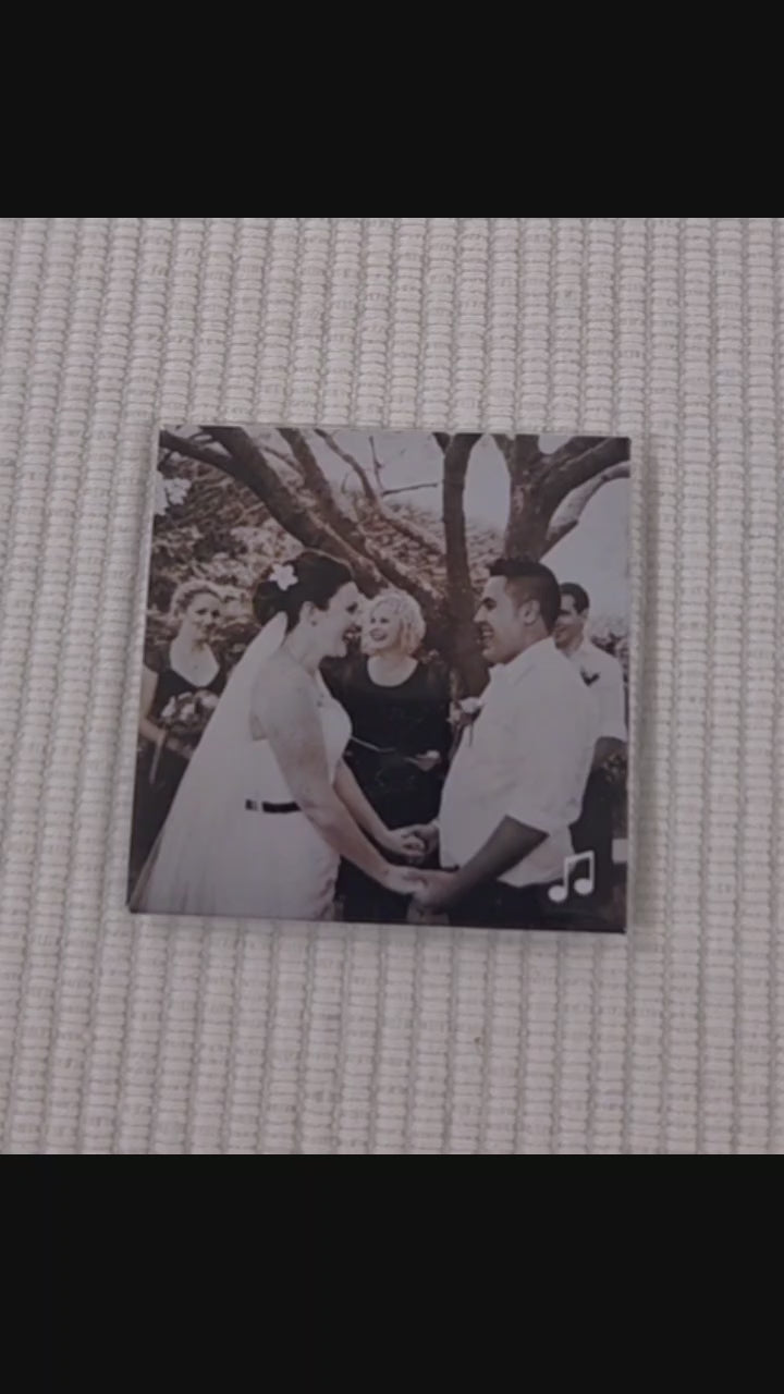 Video of a custom photo magnet which is of a couple on their wedding day. a finger points to a music logo in the bottom right corner. a phone then scans the magnet with the back of the phone and a link to a youtube video with the couple's wedding song begins to play