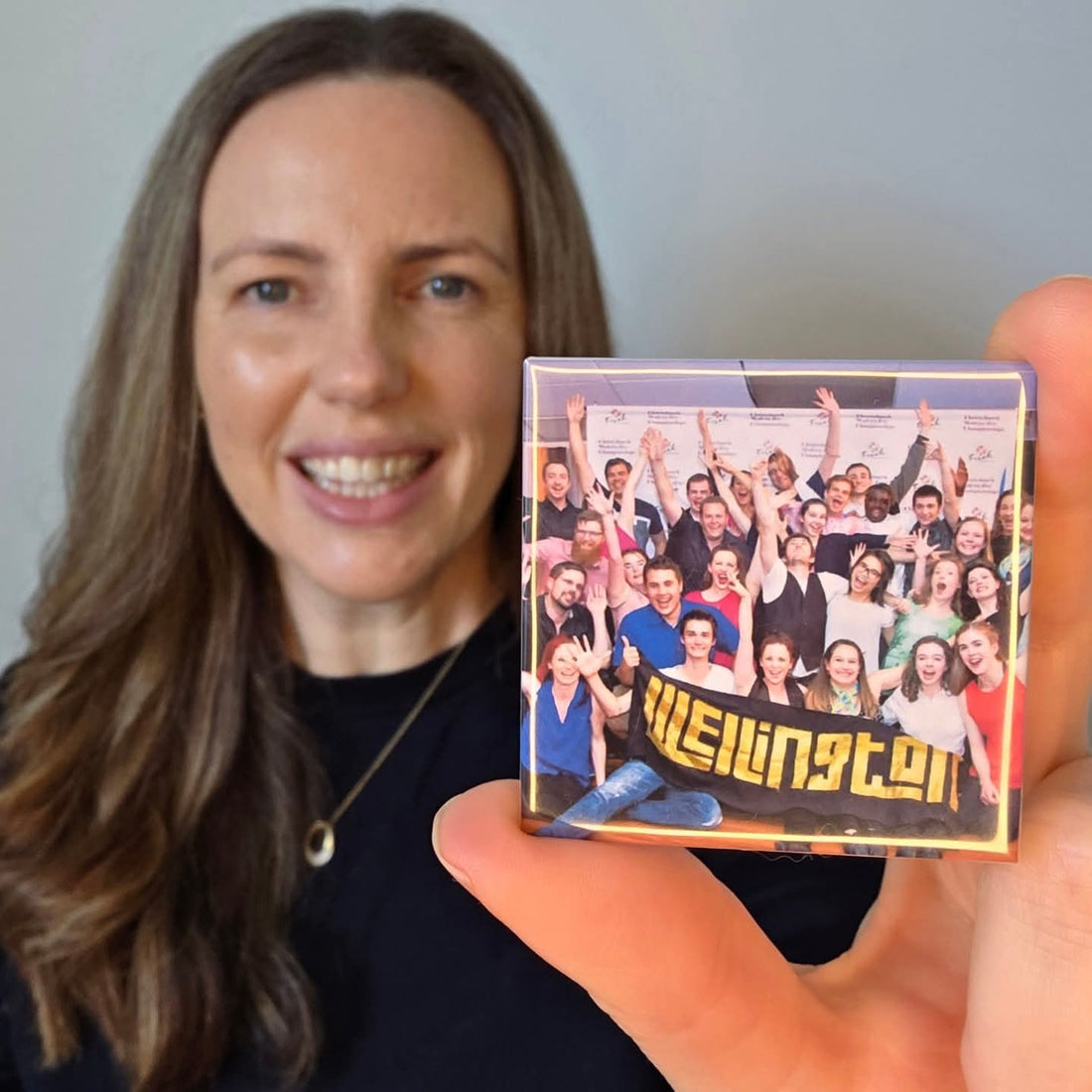 Photo of a woman with brown hair holding a 5cm x 5cm magnet towards the camera which has been made using one of her photos.  The magnet photo is of a grip of dancers gathered together at an event