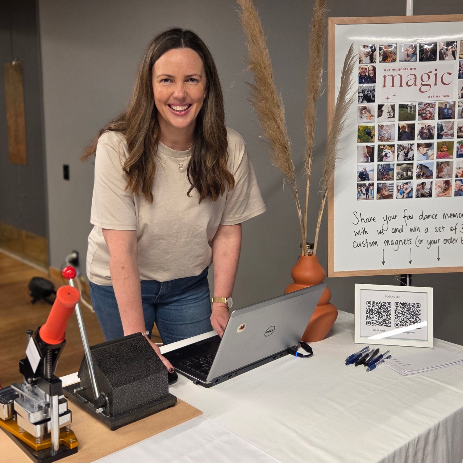 Women smiling and standing behind a market stand with magnet making equipment in front of her and a magnetic board full of pictures of custom photo magnets