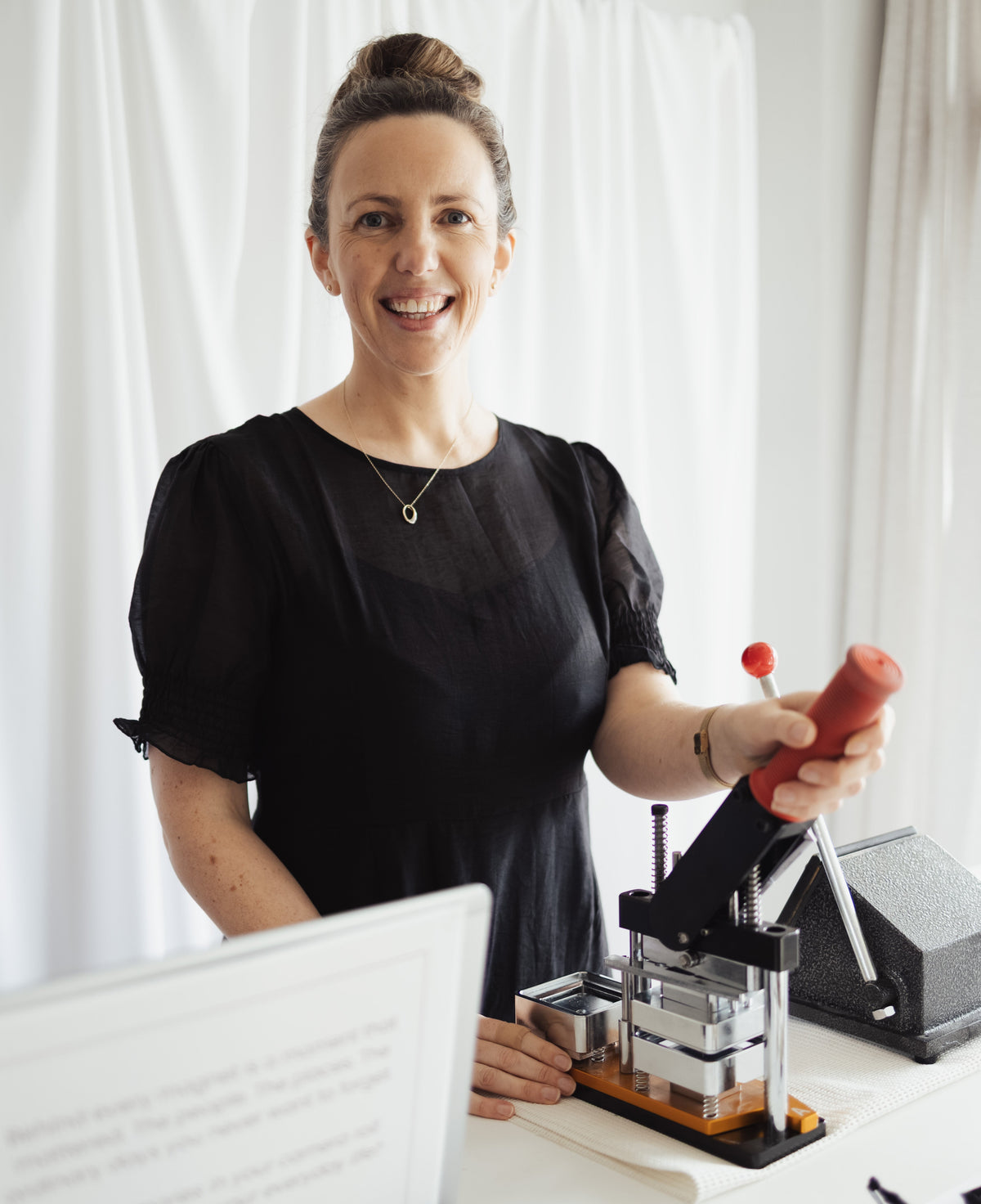 A woman in a black dress standing behind a counter with a hand-press in front of her which is used for making custom photo magnets. she is smiling and has one hand on the press